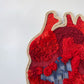 Arte textil en forma de corazón, confeccionado en tonos rojos vibrantes, destacando una variedad de texturas y patrones intrincados